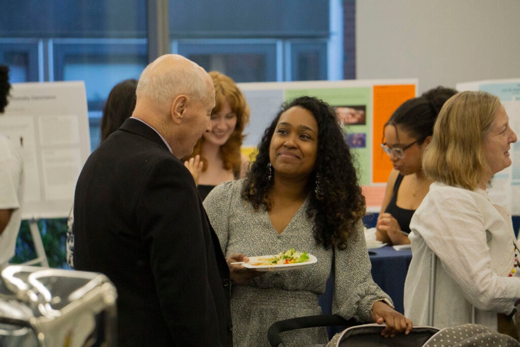 Professor Dennis McNamara speaks with Professor Alyssa Newman who holds a plate with one hand and rests the other on a baby stroller